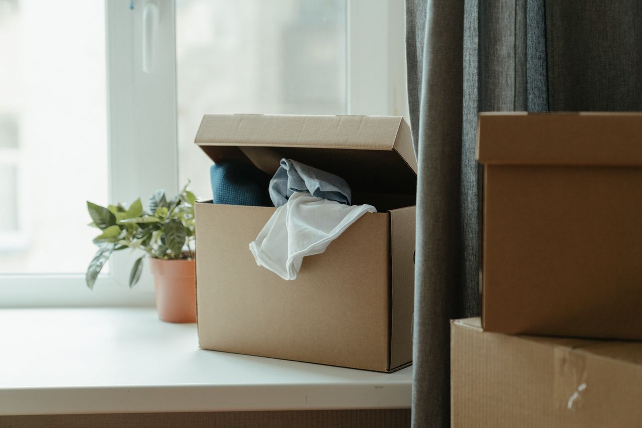 Boîte en carton ouverte avec des vêtements près d'une fenêtre, illustrant un déménagement ou une relocalisation.